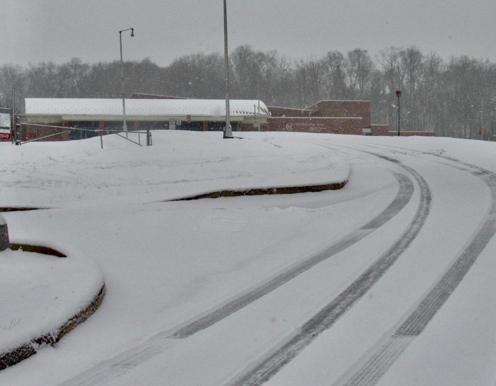 A snowy scene showing a snow-covered driveway leading to a school building. The building, identified as the Columbia Borough School Districtβs Hill Campus, is partially visible in the background with trees lining the horizon. Snow continues to fall, and tire tracks are visible on the curved road in the foreground. A streetlight and sign are present along the driveway.